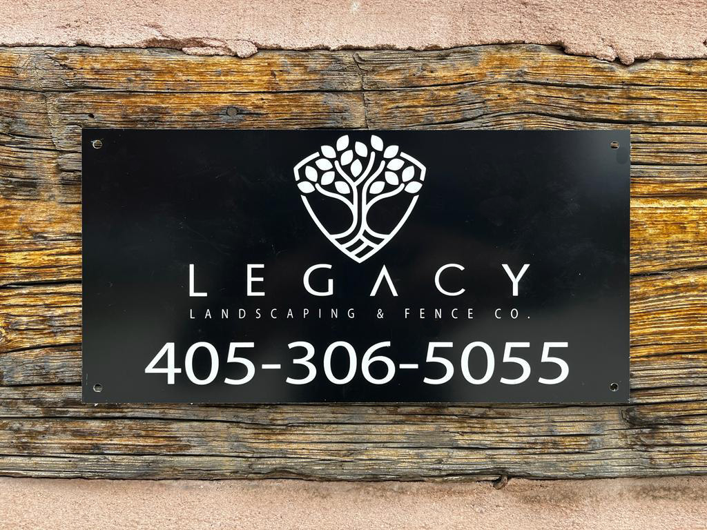 Custom black aluminum fence sign for Legacy Landscaping & Fence Co. mounted on a rustic wood plank with white logo and phone number